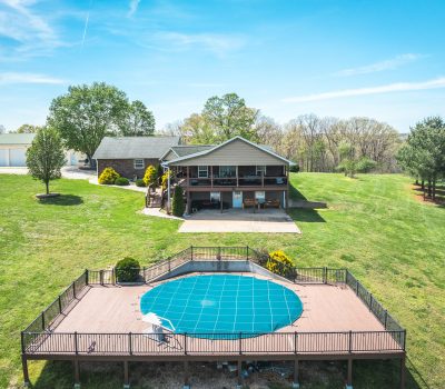 A large house with a covered porch sits in a spacious grassy yard. In the foreground, there is a round, covered swimming pool surrounded by a wooden deck and black railing under a clear blue sky.