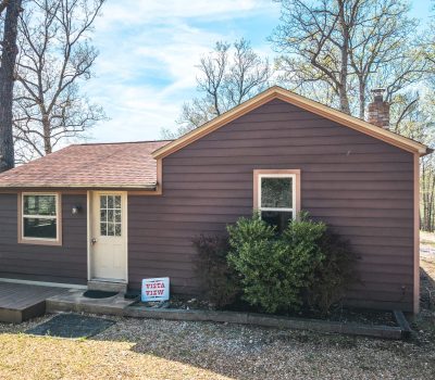 A small brown wooden cabin with a red shingle roof sits among tall trees. A sign reading "VISTA VIEW" is placed by bushes near the front door. The sky is clear and blue.