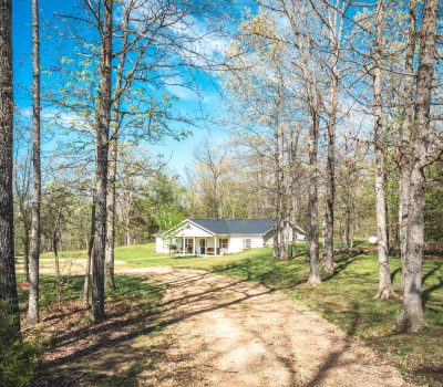 A small white house with a porch sits at the end of a dirt driveway, surrounded by tall trees with sparse green leaves on a sunny day. The sky is bright blue with a few clouds.