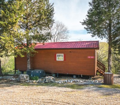 A small brown wooden cabin with a red metal roof sits among tall trees, with a gravel area and rocks in front. A single window is visible, and a picnic table is in the background.
