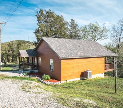 A small wooden cabin with a front porch sits on a grassy hill, surrounded by trees and shrubs, under a blue sky. A gravel driveway and utility pole are visible to the left.
