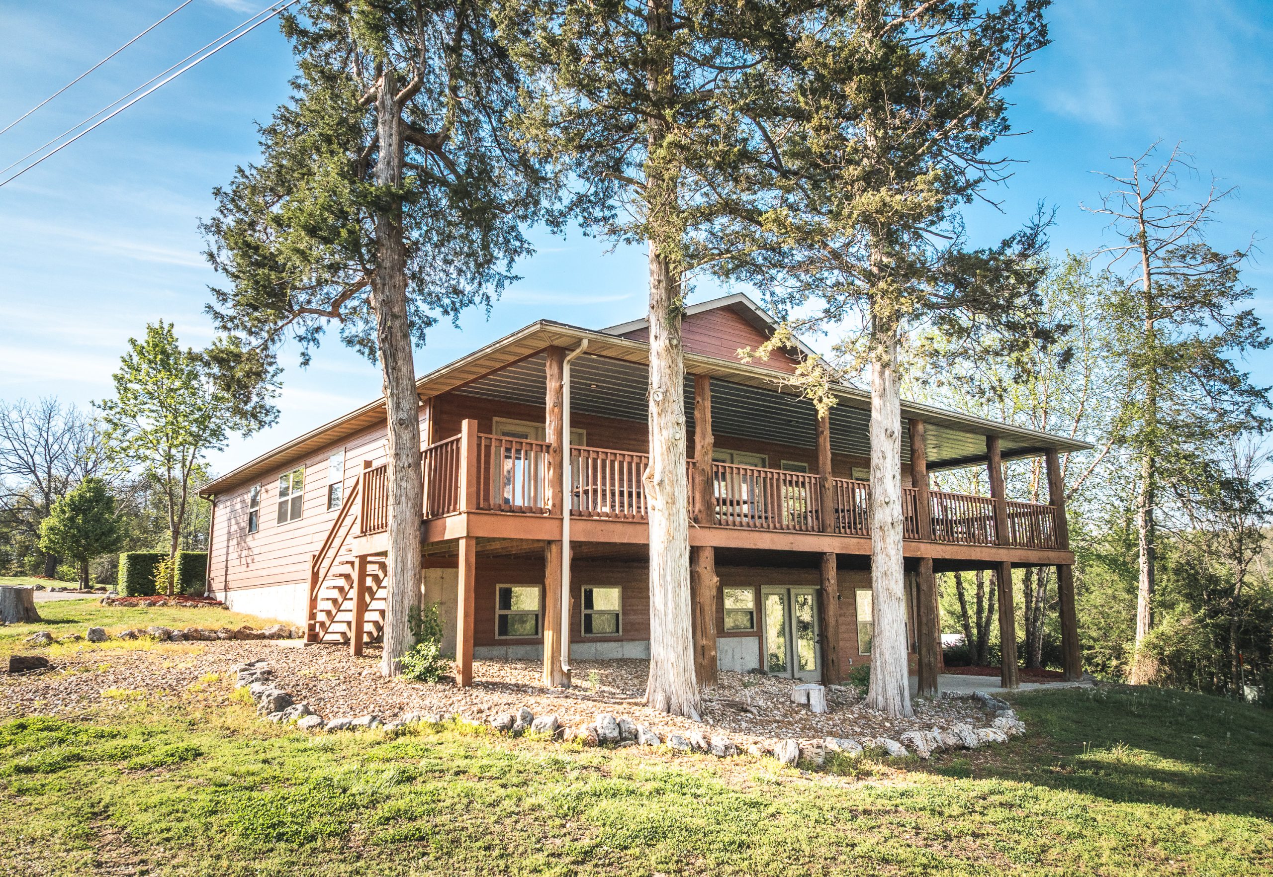 A two-story wooden house with a large wraparound porch, surrounded by tall trees and greenery, sits on a grassy lawn under a clear blue sky.