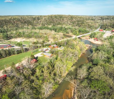Aerial view of a river winding through a green, forested landscape with scattered buildings, a bridge crossing the river, open fields, and a road leading to a small settlement under a blue sky.