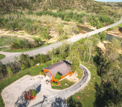 Aerial view of a red building with a curved driveway surrounded by trees, near a road and a river. A bridge crosses the river, and forested hills fill the background under a clear sky.