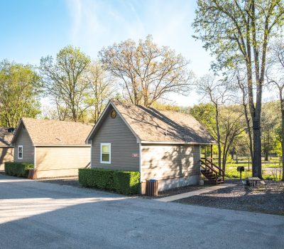 Small, beige cabins with gabled roofs stand in a row beside a paved road, surrounded by green trees and bushes on a sunny day. A picnic table and trash cans are visible near the cabins.