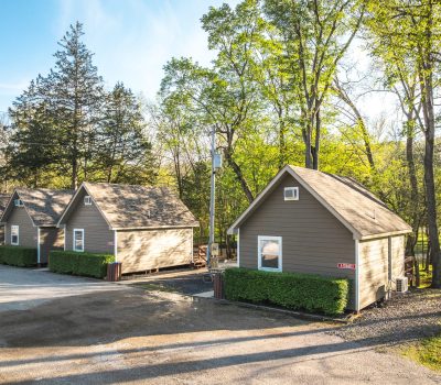 Four small brown cabins with white trim sit in a row along a gravel road, surrounded by green trees and bushes on a sunny day.