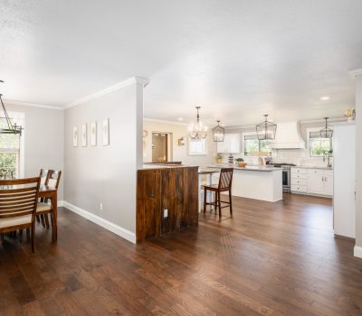 Open-concept home interior with a dining area on the left, featuring a wooden table and chairs, and a bright kitchen on the right with white cabinets, a central island, and hardwood floors throughout.