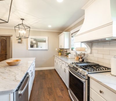 Bright, modern kitchen with white cabinets, marble countertops, stainless steel appliances, and wood flooring. Large island with a bowl, pendant lights above, and a decorative mirror on the beige wall.