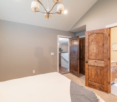 A bedroom with beige walls, a bed with white bedding, wooden double doors, an open door to a bathroom with a toilet, and a ceiling light fixture. Natural light enters from the bathroom window.