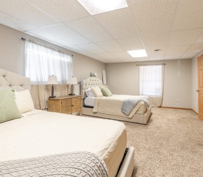 A spacious bedroom with two tufted beige beds, light bedding, a wooden nightstand with a lamp, two large windows with sheer curtains, beige carpet, and wooden double closet doors.