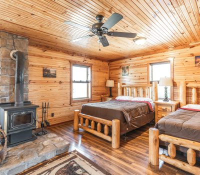 A cozy log cabin bedroom with wood-paneled walls and ceiling, two double beds with log frames, a wood-burning stove on a stone hearth, and rustic decor throughout. Natural light enters through multiple windows.