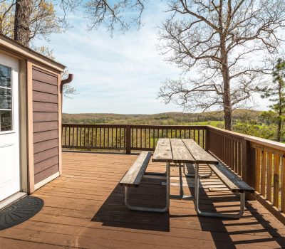 A wooden deck with a picnic table overlooks a scenic landscape of rolling hills and trees. The deck is attached to a building with brown siding and a white door. The sky is clear and sunny.