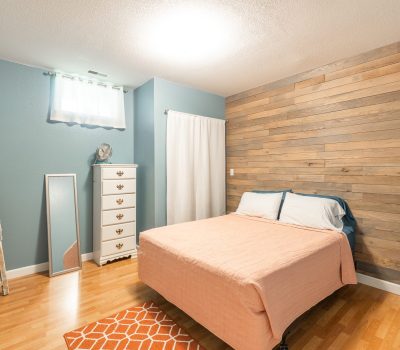 A cozy bedroom with a peach bedspread, wooden accent wall, blue walls, and light wood floors. There’s a white dresser, ladder shelf with towels, mirror, orange rug, and small lamp on a wall shelf.