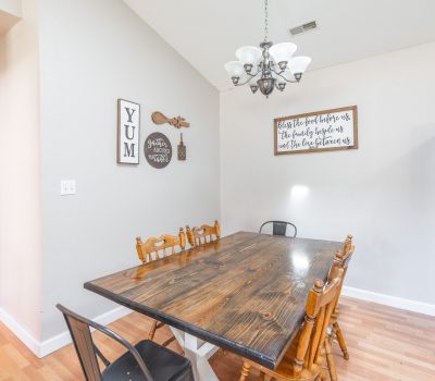 A wooden dining table with mixed chairs sits under a chandelier in a bright room with light walls and hardwood floor. Wall art and a silver refrigerator are visible in the background.