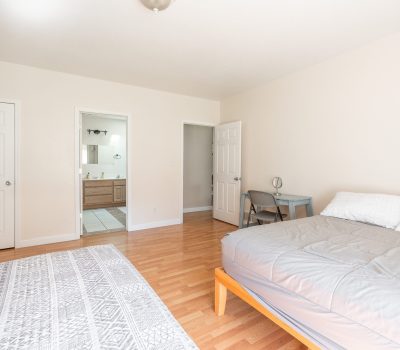 A bright, minimal bedroom with light wood floors, a double bed with gray bedding, a small desk and chair, a white patterned rug, and an open door leading to a bathroom with a double vanity.