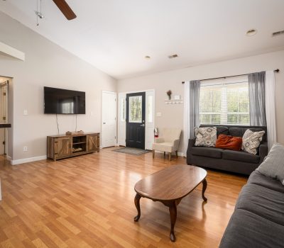A cozy living room with wood floors, two gray sofas with patterned pillows, a wooden coffee table, a TV mounted on the wall, large window with curtains, and a black front door letting in natural light.