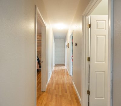 A well-lit hallway with light-colored walls, wooden flooring, and several open doors leading to different rooms. Natural light is visible at the end of the hallway.