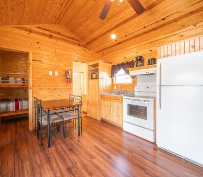 Cozy wooden cabin interior with a small dining table, white refrigerator, stove, microwave, and coffee maker. A bunk bed is visible in a side room, and natural light comes through a window with dark curtains.