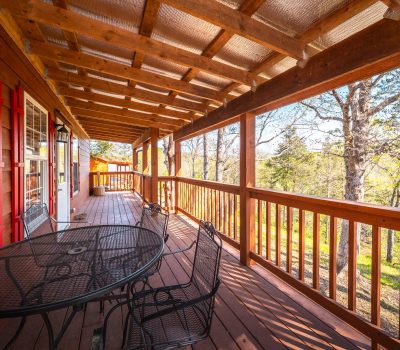 A wooden porch with a sloped roof, black metal table and chairs, and red shutters overlooks a sunny, tree-filled yard. Sunlight filters through the trees, casting shadows on the wooden floor.