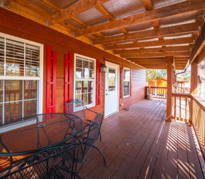 A wooden porch with a slatted roof, red shutters, large windows, and a white door. There is a black metal patio table with chairs, and the porch is surrounded by trees and sunlight.