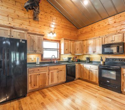 Rustic kitchen with wood-paneled walls and cabinets, black appliances including fridge, oven, microwave, and dishwasher, hardwood floors, and a window over the sink letting in natural light.