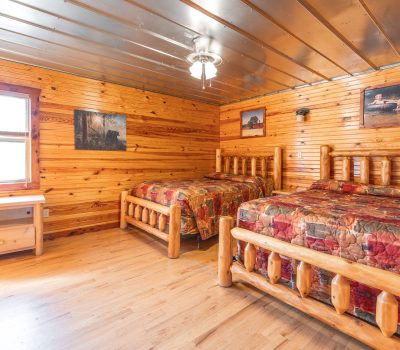 A cozy log cabin bedroom with wooden walls and ceiling, two rustic wooden beds with patchwork quilts, a wooden desk by the window, and nature-themed paintings on the walls.
