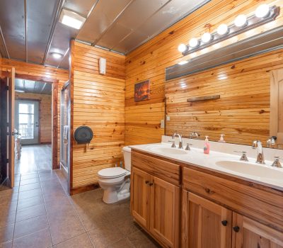 A rustic bathroom with wood-paneled walls, a double sink vanity, large mirror with lights above, a toilet, tile flooring, and a hallway leading to a room with natural light.