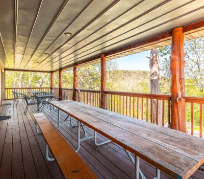 A covered wooden porch with long picnic tables and chairs overlooks trees and distant hills, with sunlight streaming through the railing.