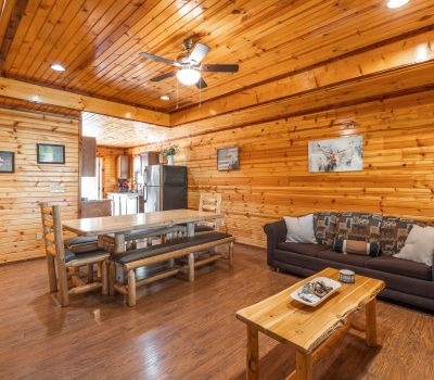 A cozy wooden cabin interior features a living area with two couches, a coffee table, and a dining table with benches. The kitchen with stainless steel appliances is visible in the background, and wood paneling covers the walls and ceiling.