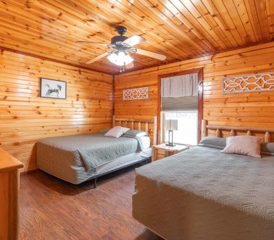 A cozy wooden cabin bedroom with two double beds, gray bedspreads, a wooden dresser, a ceiling fan, wall decor, and a window letting in natural light. The room features wood-paneled walls and ceiling.