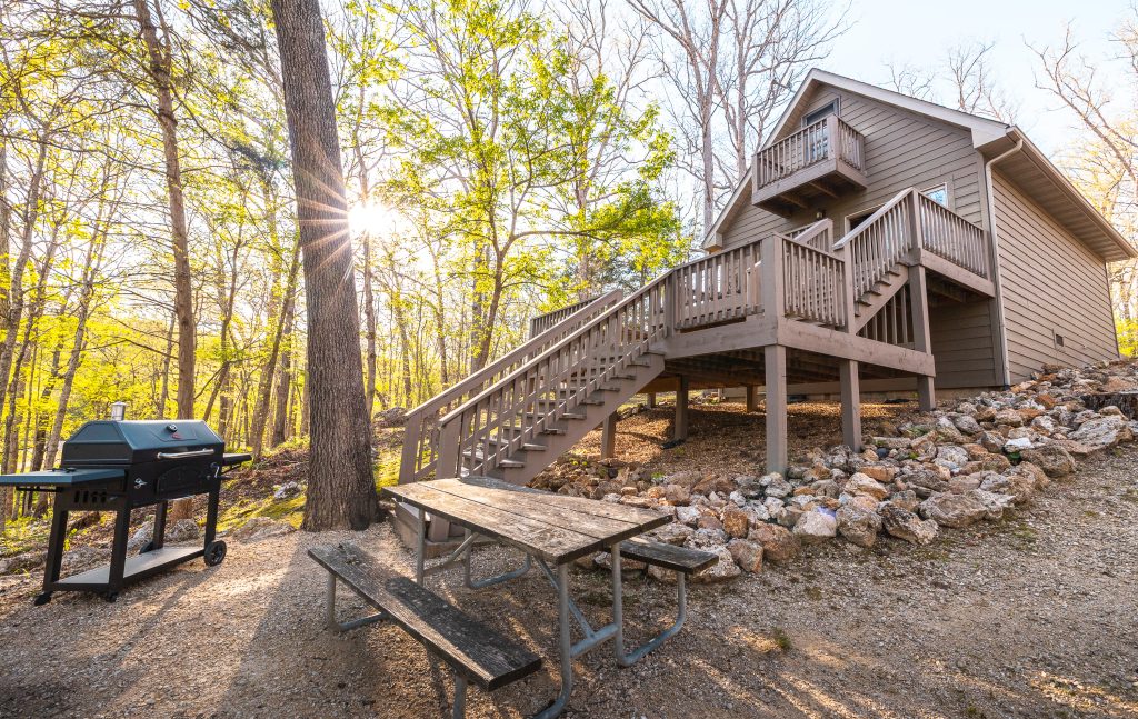 A wooden cabin with an elevated deck sits among trees and rocks. A picnic table and a barbecue grill are nearby, with sunlight streaming through the forest in the background.
