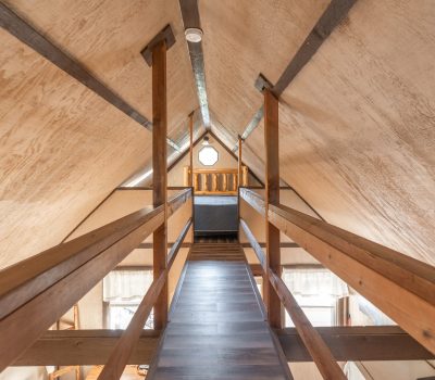 A narrow wooden loft walkway with railings leads to a small bed placed against the far wall, beneath a triangular window in a cozy attic room with slanted ceilings and exposed wooden beams.
