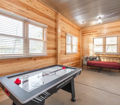 A room with wood-paneled walls features an air hockey table in the foreground and a daybed with a red blanket in the back. Sunlight comes through multiple windows, and cleaning tools are visible in the corner.