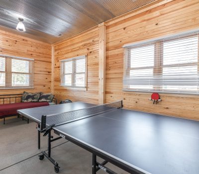 A room with wood-paneled walls, large windows with blinds, a black ping pong table, a paddle and ball on the windowsill, and a daybed with pillows and a red cover in the corner.