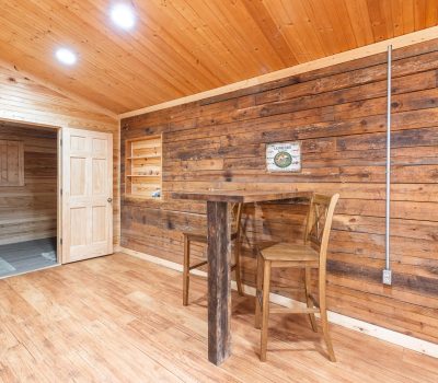A cozy room with wooden walls, ceiling, and floor features a rustic high table with two chairs near a wall, open shelves, a door to another room, and a small metal sign as decoration. The lighting is warm and inviting.