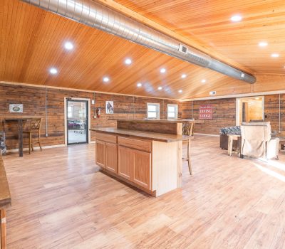 Spacious rustic kitchen and living area with wood floors, wood-paneled walls and ceiling, large central island, dining table, cozy seating, and exposed air duct, brightly lit by ceiling lights and natural sunlight.