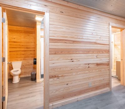 A hallway with light wood-paneled walls and gray flooring. Two open doors reveal a bathroom with a toilet and another room with a sink. A fire extinguisher is mounted on the wall near the doors.