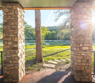 View from a shaded porch with stone columns, looking out onto a grassy park with trees and a dirt path, bathed in bright sunlight with sun rays streaming in from the right.