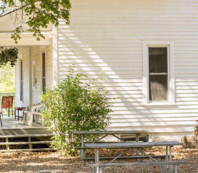 A quaint white wooden house with a wraparound porch featuring two red chairs and a hanging basket. A bush grows beside the house, and in the foreground, there are picnic tables on gravel under the shade of a tree. Sunlight creates patterns on the siding.