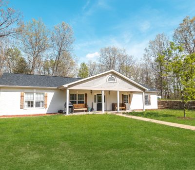 Single-story white house with a dark roof, wooden shutters, and a covered front porch. The house sits on a grassy lawn with a paved walkway, surrounded by trees and a stone wall under a blue sky.