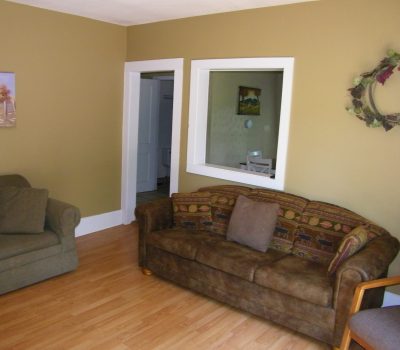 A cozy living room with tan walls and light wooden flooring features a brown two-seater sofa with patterned cushions, a green armchair, and a wooden chair with a cushion. There is a landscape painting on the wall, a wreath, and a large window opening to another room.